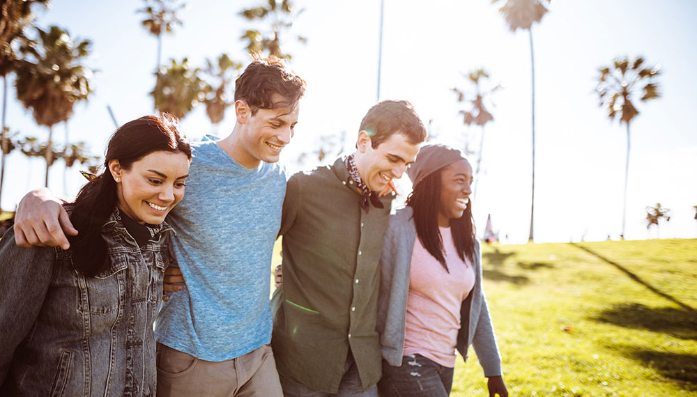 4 friends, 2 men and 2 women, with arms over shoulders walking outside with palm trees and sun in the background
